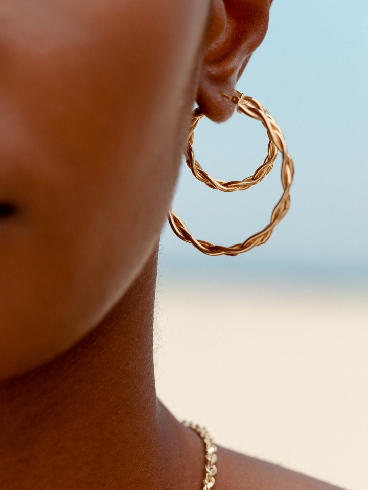 Close up shot of models ear wearing the Product shot of the Braided Hoops (Vermeil Braided Hoops Diameter: 40mm Thickness: 4mm) along with the Mini Braided Hoops (Vermeil Braided Hoops Diameter: 24.6mm Thickness: 4mm) Background: Blue sky 